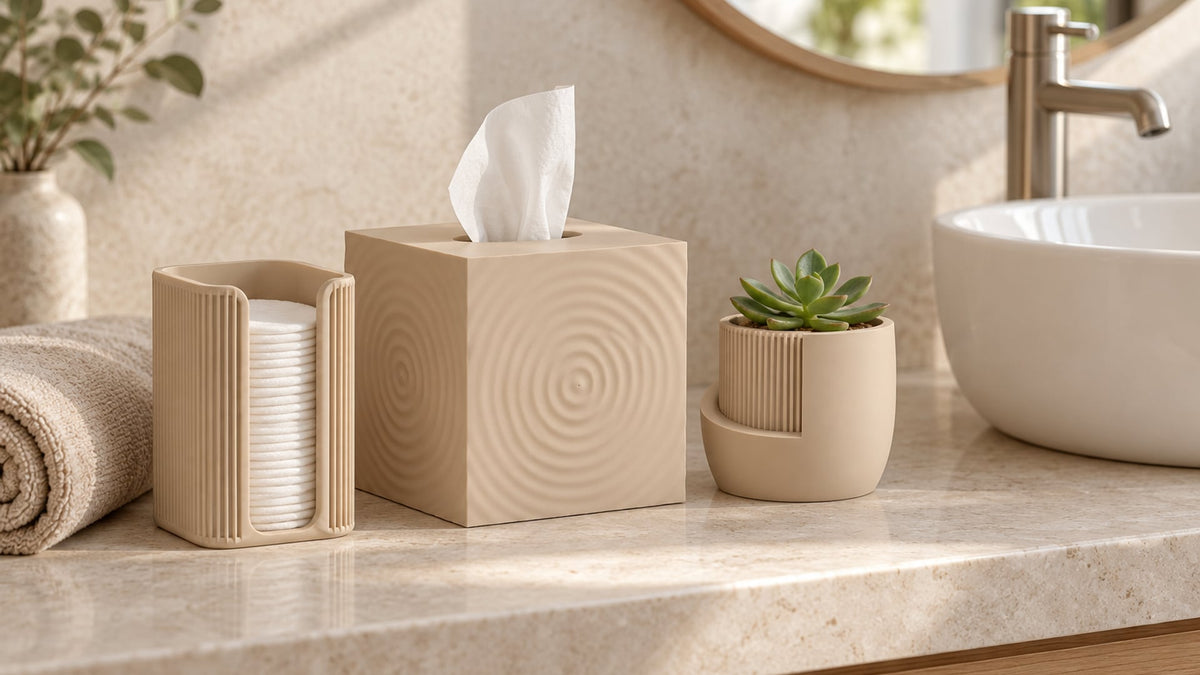 Beige ribbed cotton pad holder with cotton pads, a cube tissue box cover with circle relief and a small japandi plant pot with a succulent on a light stone bathroom vanity next to a washbasin and a rolled towel