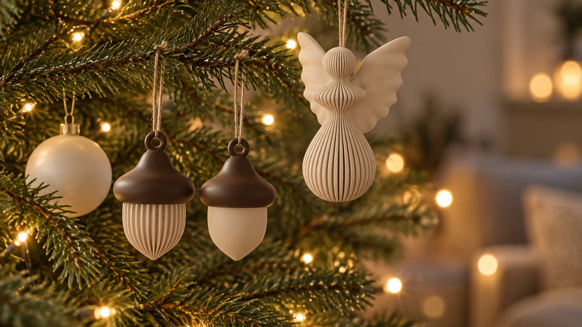 Close-up of a real spruce branch with warm LED fairy-light bokeh: two small 3D-printed acorn ornaments (beige with chocolate caps) and a beige ribbed angel hang from the twigs on natural twine