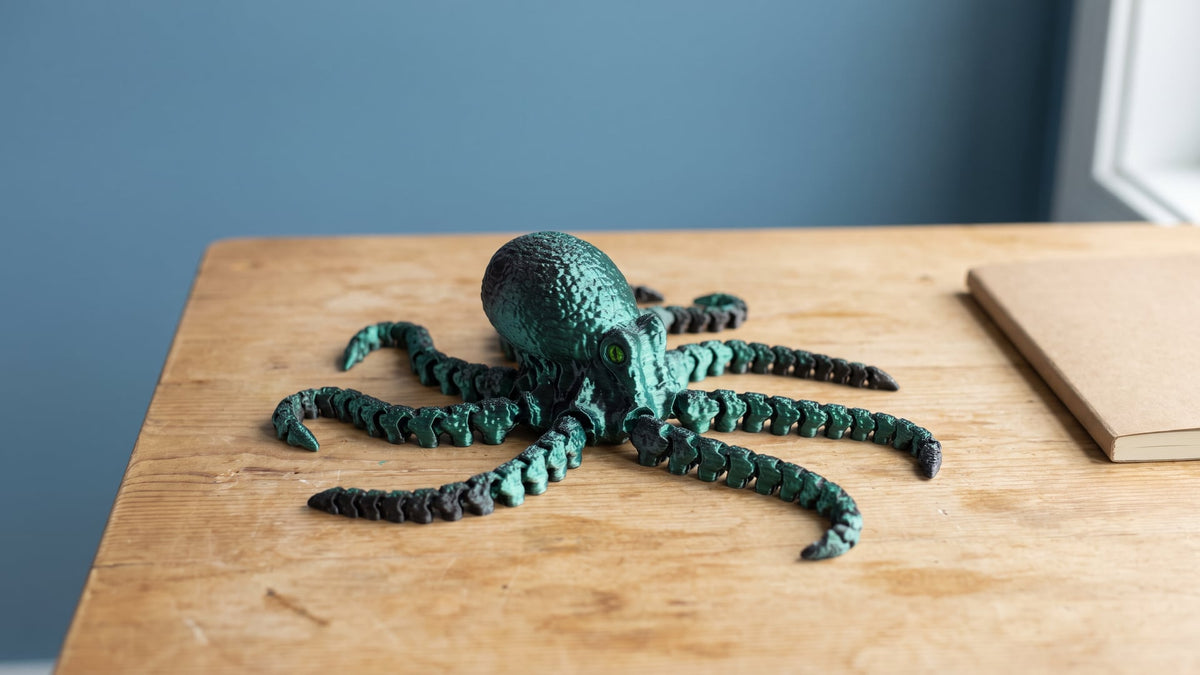 A black and teal-green articulated 3D-printed flexi octopus on a light wooden desk, its eight segmented arms spread out in a star, against a calm blue wall with a plain notebook at the edge.
