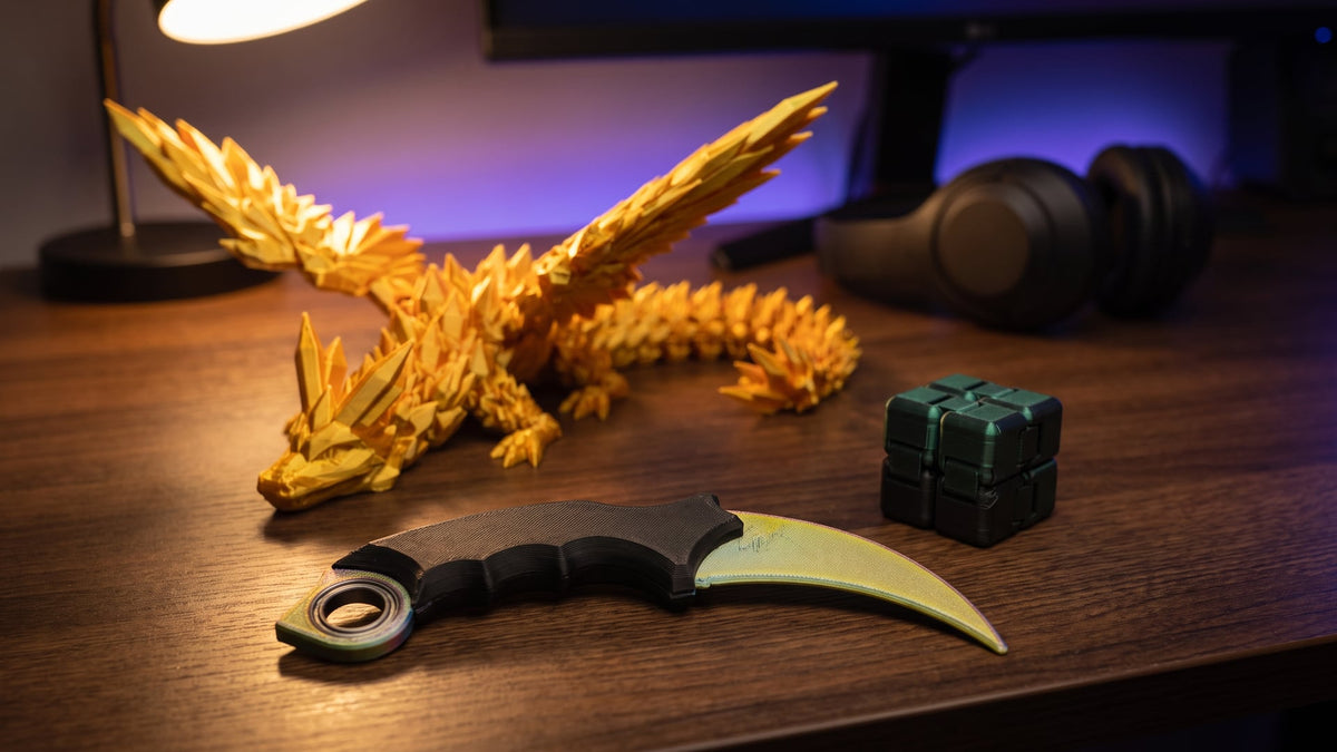 Gold 3D printed winged dragon, a karambit fidget spinner with a black grip and a black-green infinity cube on a wooden desk in lamplight, headphones in the background