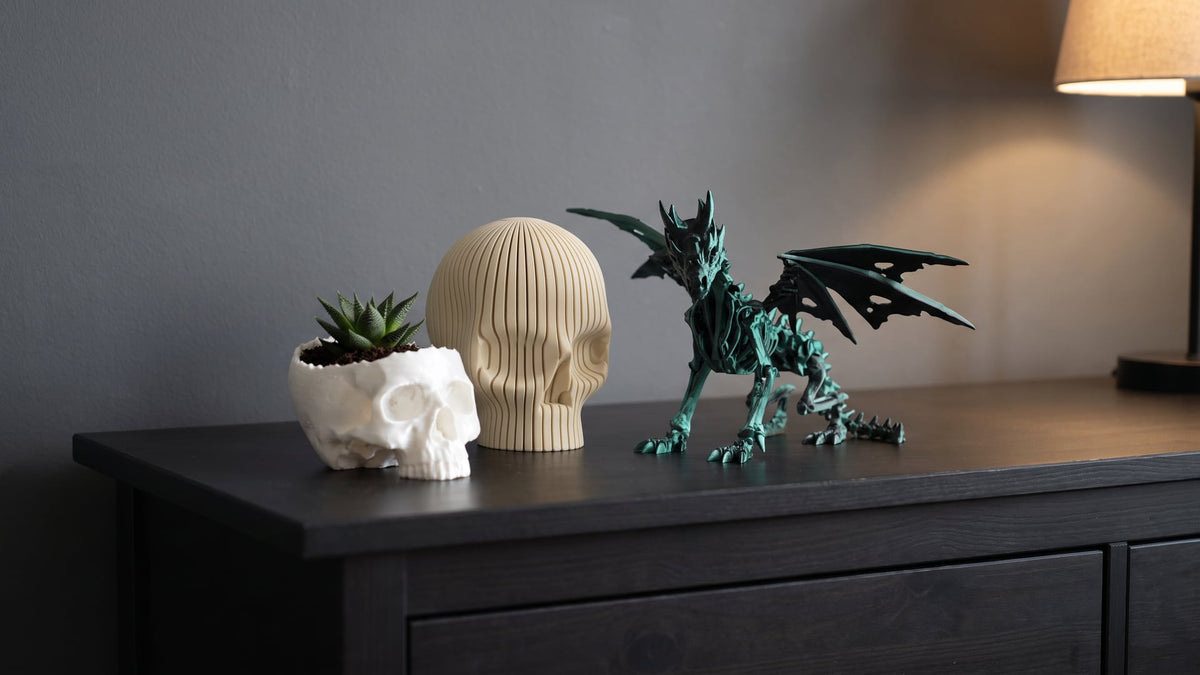 Dark wooden chest of drawers by a grey wall: a white skull planter with a succulent, a beige ribbed decorative skull and a black-green articulated skeleton dragon with spread wings, lit from the side by a table lamp