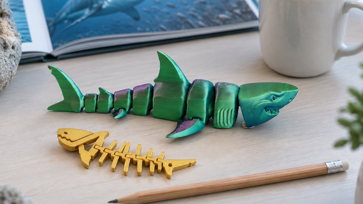 Articulated 3D-printed great white shark in green-purple silk curved as if swimming on a light desk, a gold shark-skeleton keychain and a pencil beside it, a mug and an open ocean book behind